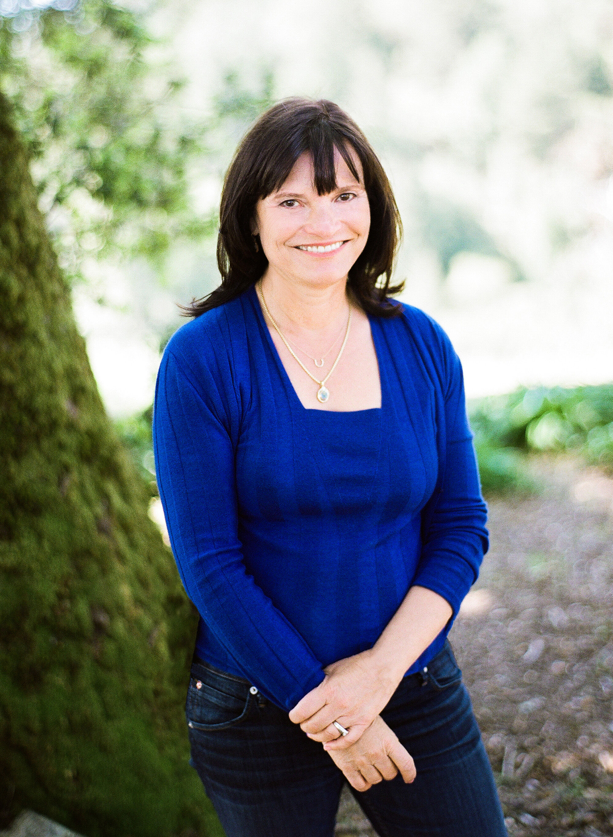 A woman with brown hair in a dark blue shirt smiling at the camera leaning on a tree