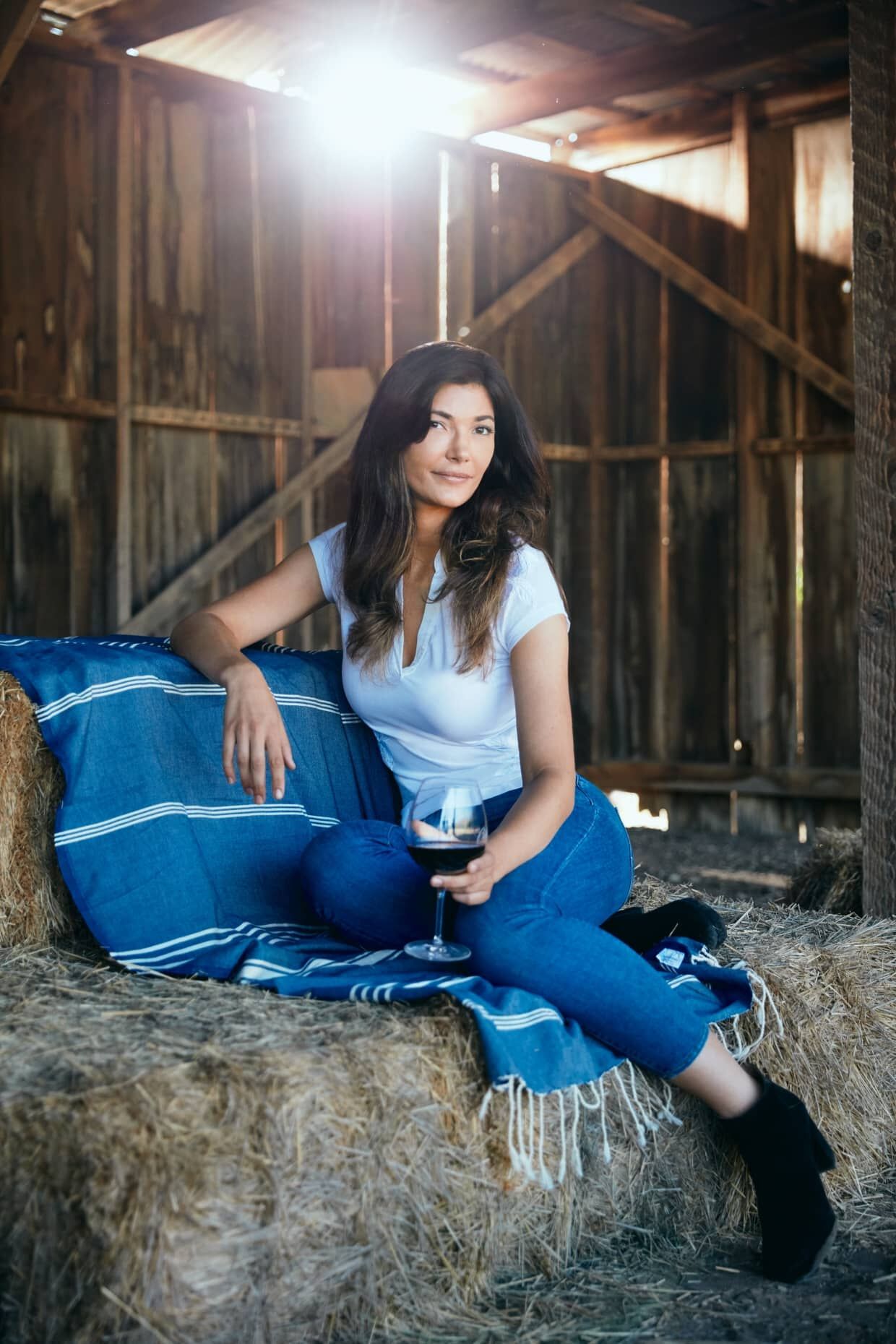 A woman in blue jeans and a white shirt sitting on a hay bale, looking at the camera with a glass of wine in hand