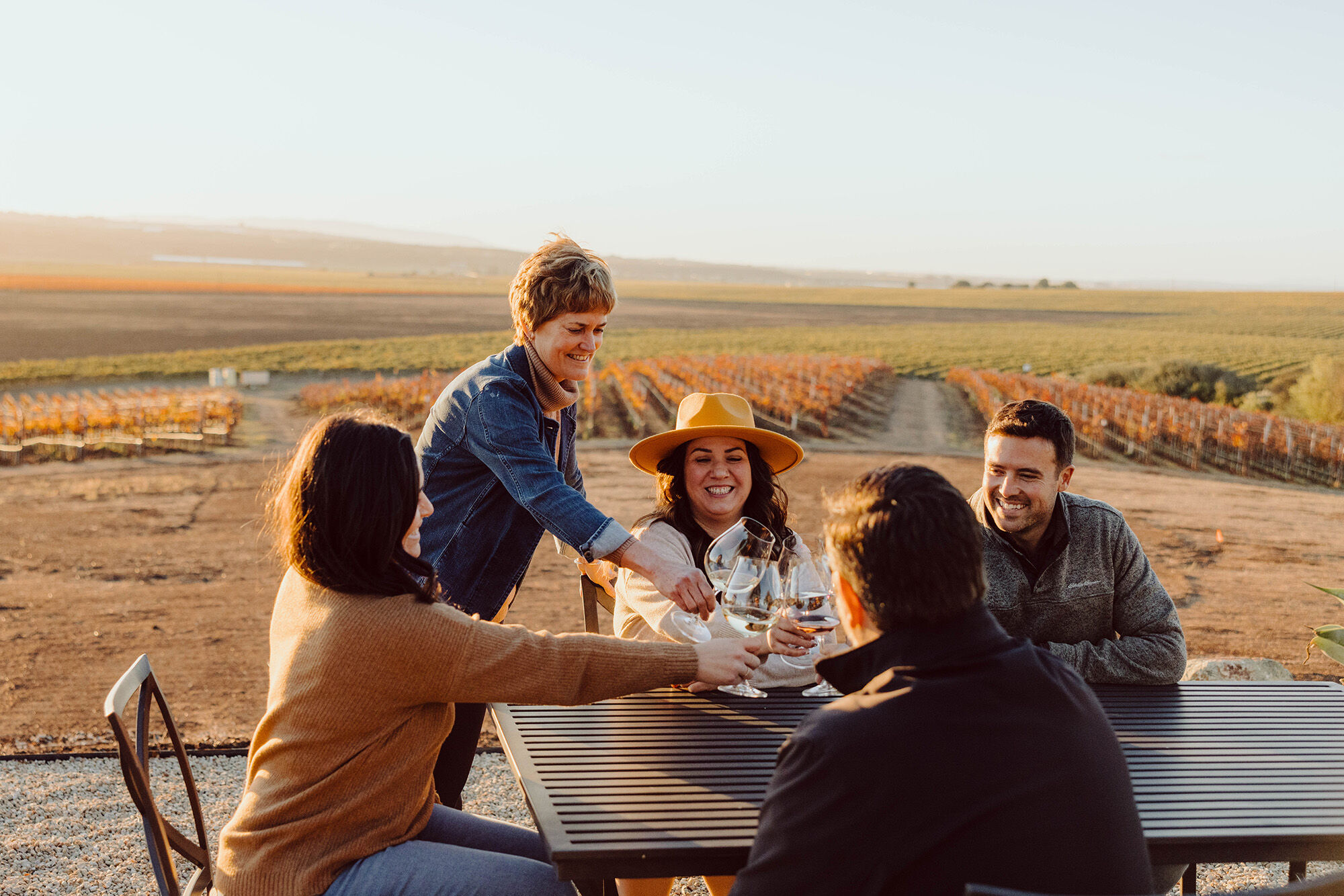 A group of friends at a table outdoor, smiling and clinking glasses with green and gold vineyards in the background.