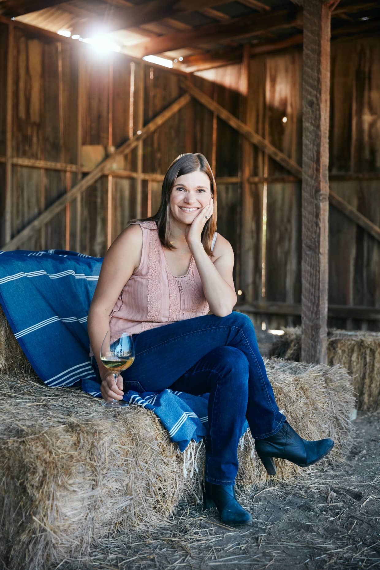 A woman in blue jeans and a pink tank top sitting on a hay bale, smiling at the camera